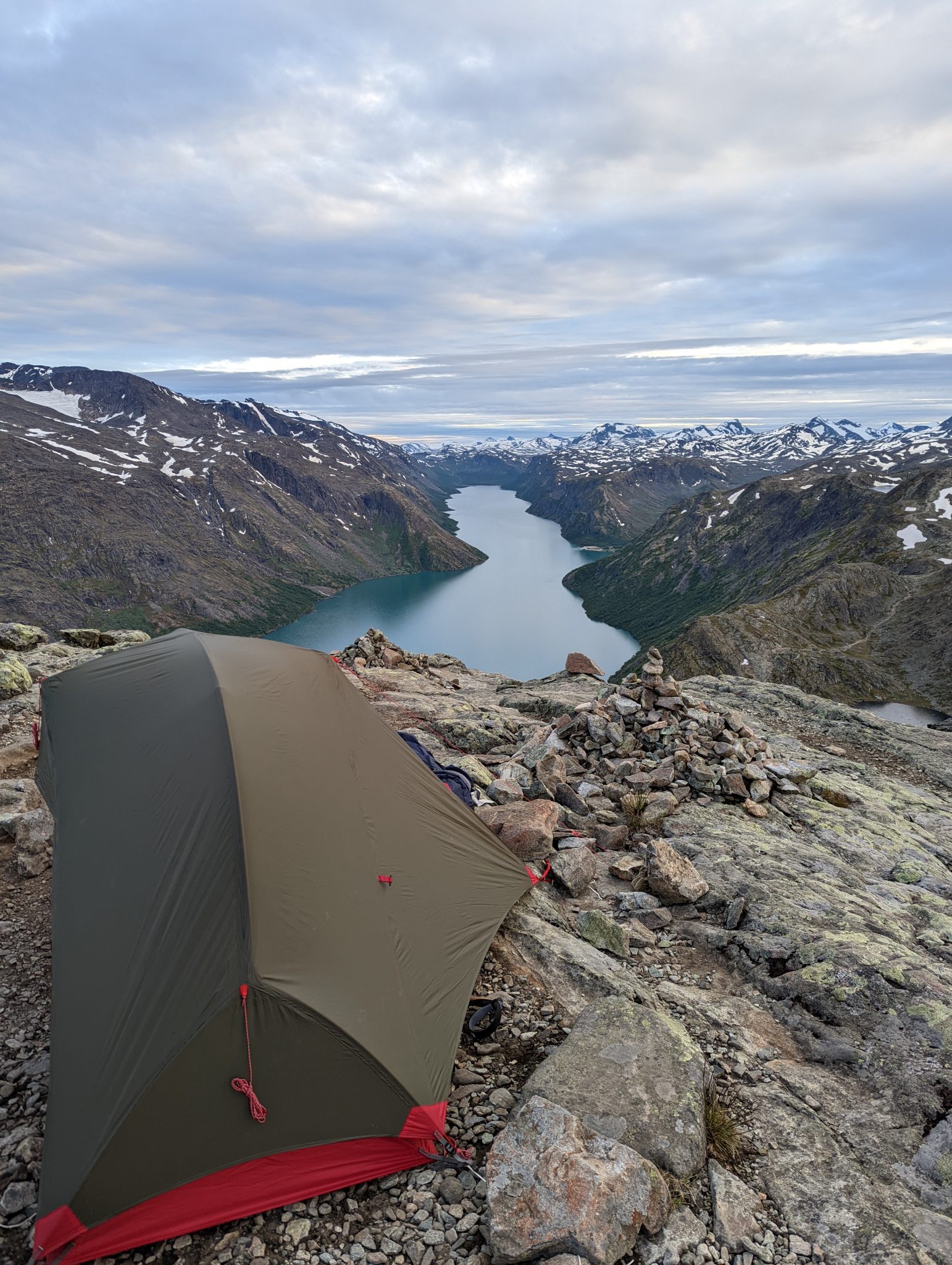 A tent on top of a mountain overlooking a fjord with snow capped mountains in the backgroud